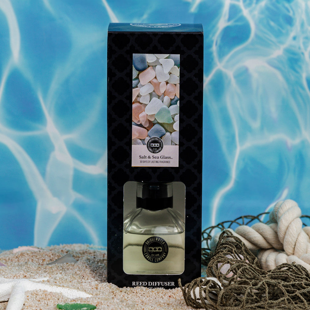 A Bridgewater Candle Company Salt & Sea Glass Reed Diffuser rests on sandy seashells, with a swimming pool and sea glass photo in the background, evoking a refreshing home fragrance.