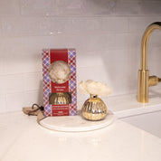 The Bridgewater Candle Company Welcome Home Mercury Flower Diffuser, featuring a cream flower top and gold vase, sits on a tray by its red plaid box, adding fragrance and elegance to the white kitchen counter near the gold faucet.