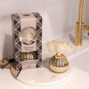 A boxed Bridgewater Candle Company Afternoon Retreat Mercury Flower Diffuser sits by an open gold vase diffuser with a cream flower on a marble tray near a gold faucet, bringing elegant fragrance to this modern bathroom.