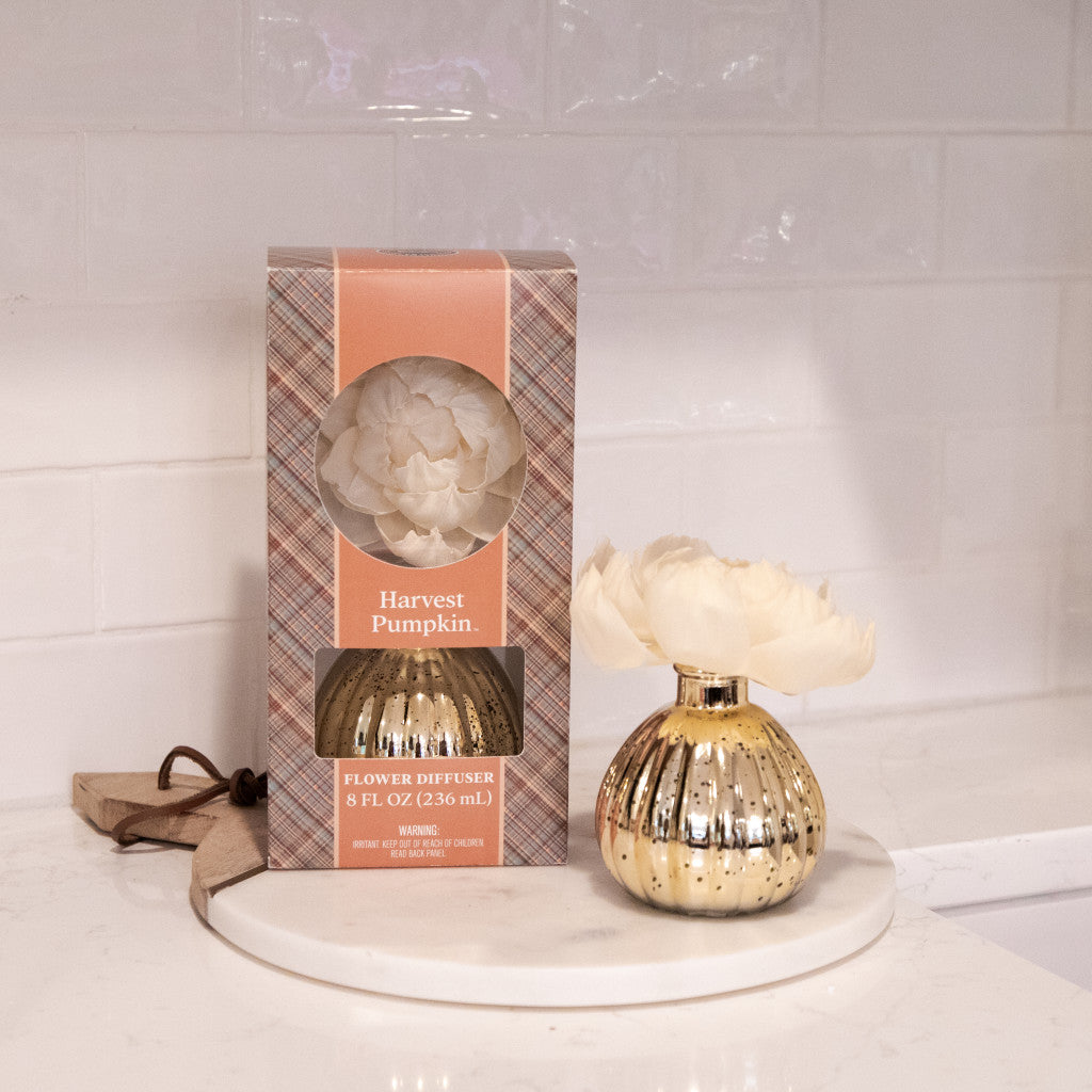 A Bridgewater Candle Company Harvest Pumpkin Mercury Flower Diffuser, featuring a pumpkin spice fragrance in a gold mercury glass vase, is displayed on a marble tray next to its box atop a white kitchen counter with a subway tile backsplash.