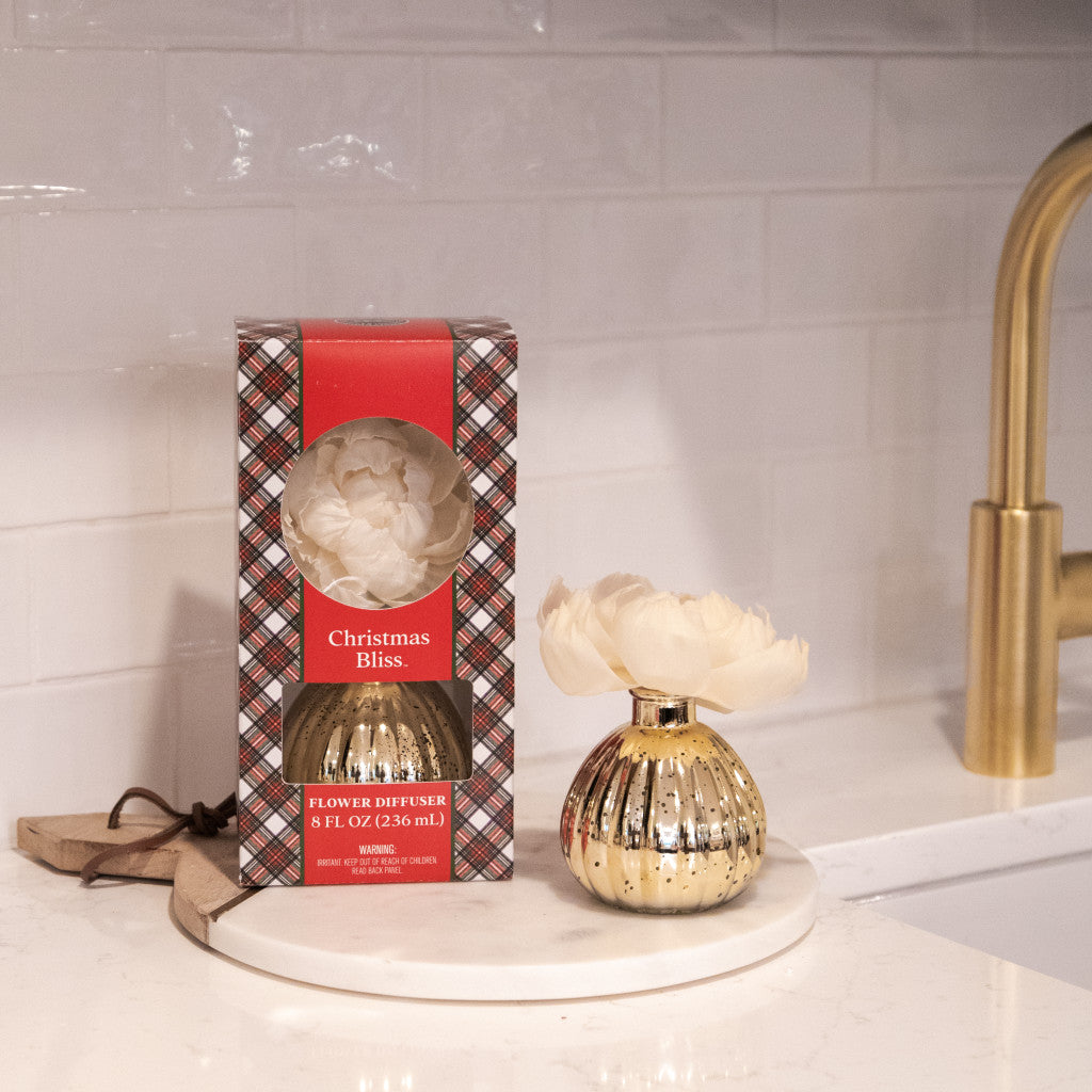The Bridgewater Candle Company Christmas Bliss Holiday Mercury Flower Diffuser, with a white flower, sits on a round marble tray beside its plaid box on a kitchen counter with a white tile backsplash and gold faucet.