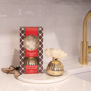 The Bridgewater Candle Company Christmas Bliss Holiday Mercury Flower Diffuser, with a white flower, sits on a round marble tray beside its plaid box on a kitchen counter with a white tile backsplash and gold faucet.