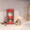 The Bridgewater Candle Company Christmas Bliss Holiday Mercury Flower Diffuser, with a white flower, sits on a round marble tray beside its plaid box on a kitchen counter with a white tile backsplash and gold faucet.