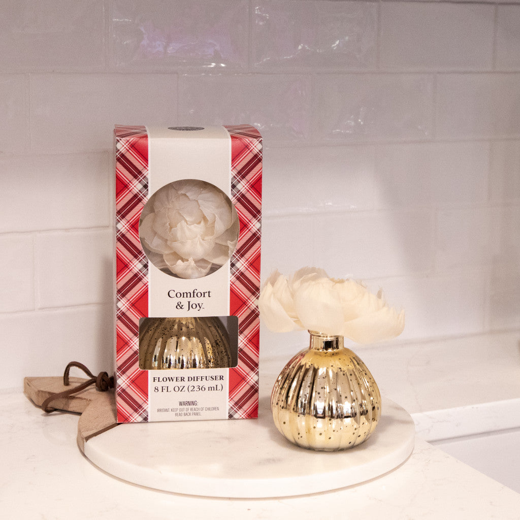 A gold Comfort & Joy Holiday Mercury Flower Diffuser by Bridgewater Candle Company, featuring a cream artificial flower, sits on a white tray beside its plaid packaging on a white countertop with a tiled backsplash.