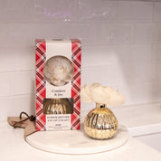 A gold Comfort & Joy Holiday Mercury Flower Diffuser by Bridgewater Candle Company, featuring a cream artificial flower, sits on a white tray beside its plaid packaging on a white countertop with a tiled backsplash.