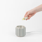 A hand pours Bridgewater Candle Company’s Sweet Grace Home Fragrance Oil into a gray ceramic electric diffuser, creating a safe, flameless scent experience against a white background.