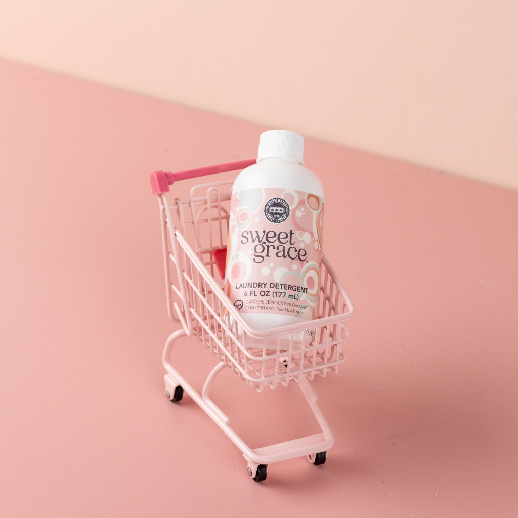 A small pink shopping cart holds a bottle of Bridgewater Candle Company’s Sweet Grace 6oz Laundry Detergent, celebrated for its fabric-safe formula and enduring scent, set against a pink background.