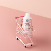 A small pink shopping cart holds a bottle of Bridgewater Candle Company’s Sweet Grace 6oz Laundry Detergent, celebrated for its fabric-safe formula and enduring scent, set against a pink background.