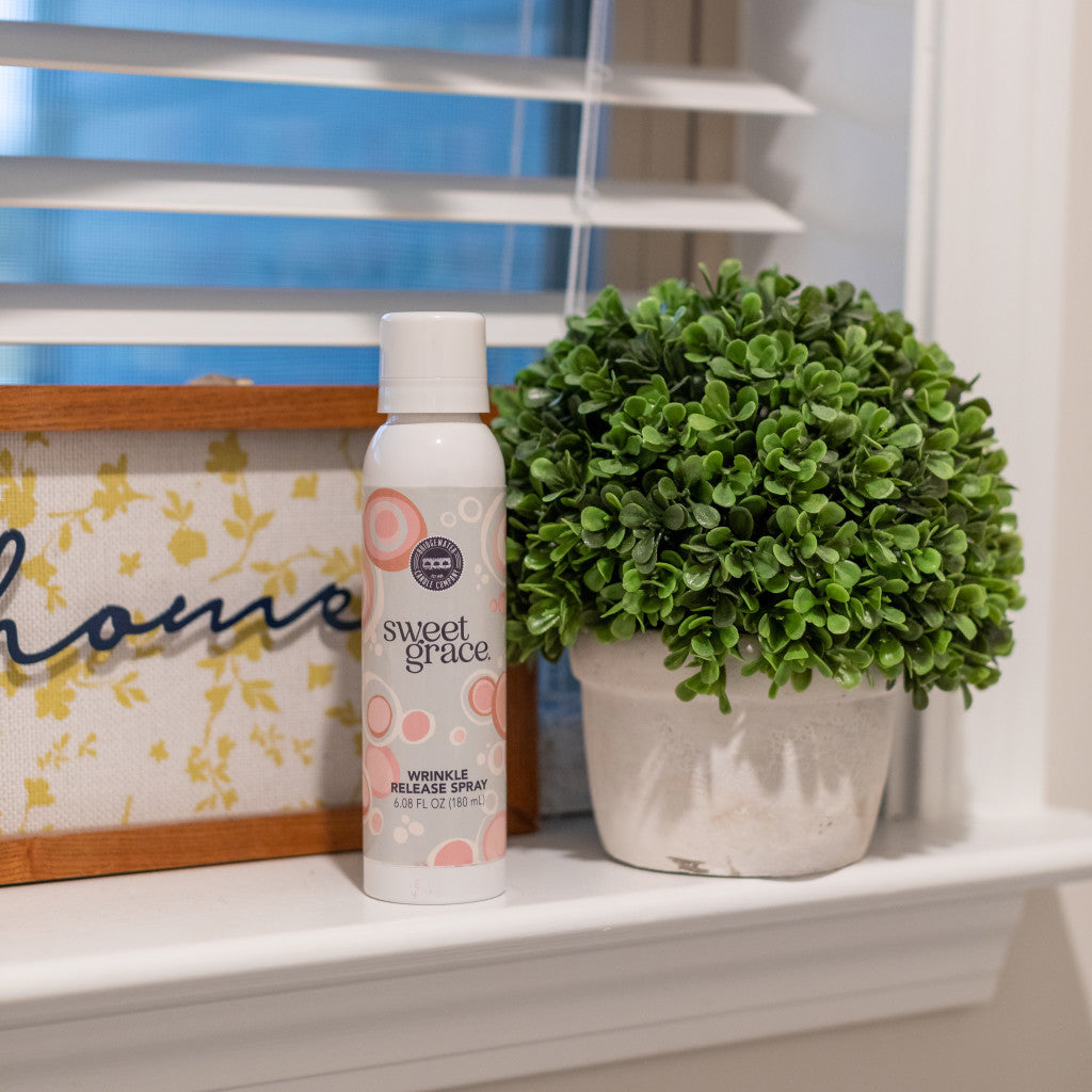 A can of Bridgewater Candle Companys Sweet Grace Wrinkle Release Spray rests on a white windowsill beside a potted plant and a framed sign with cursive text, with window blinds behind showcasing this fresh clothing spray.