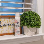A can of Bridgewater Candle Companys Sweet Grace Wrinkle Release Spray rests on a white windowsill beside a potted plant and a framed sign with cursive text, with window blinds behind showcasing this fresh clothing spray.