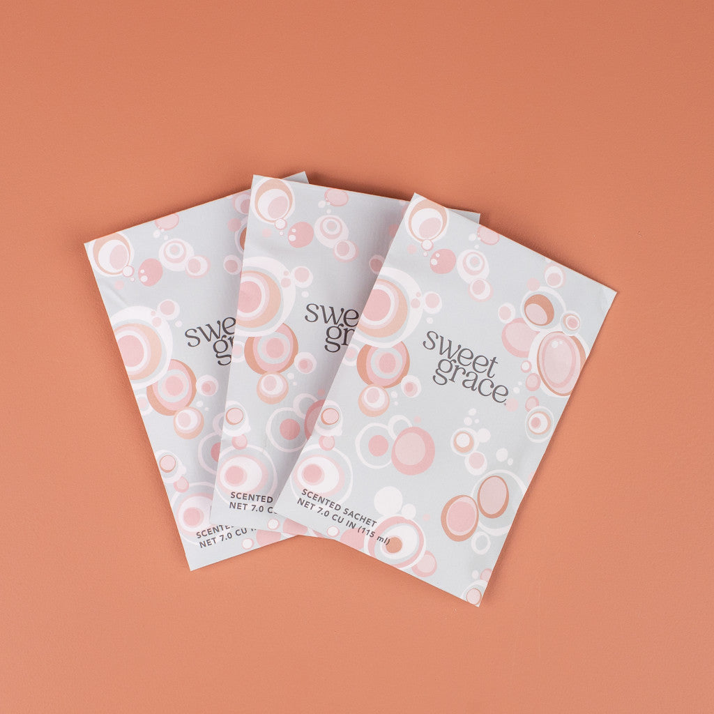 Three Bridgewater Candle Company Sweet Grace Modern Bubble Sachets, featuring pink and white bubble designs, are arranged in a fan shape on a peach background—ideal for adding a delightful fragrance to your home.