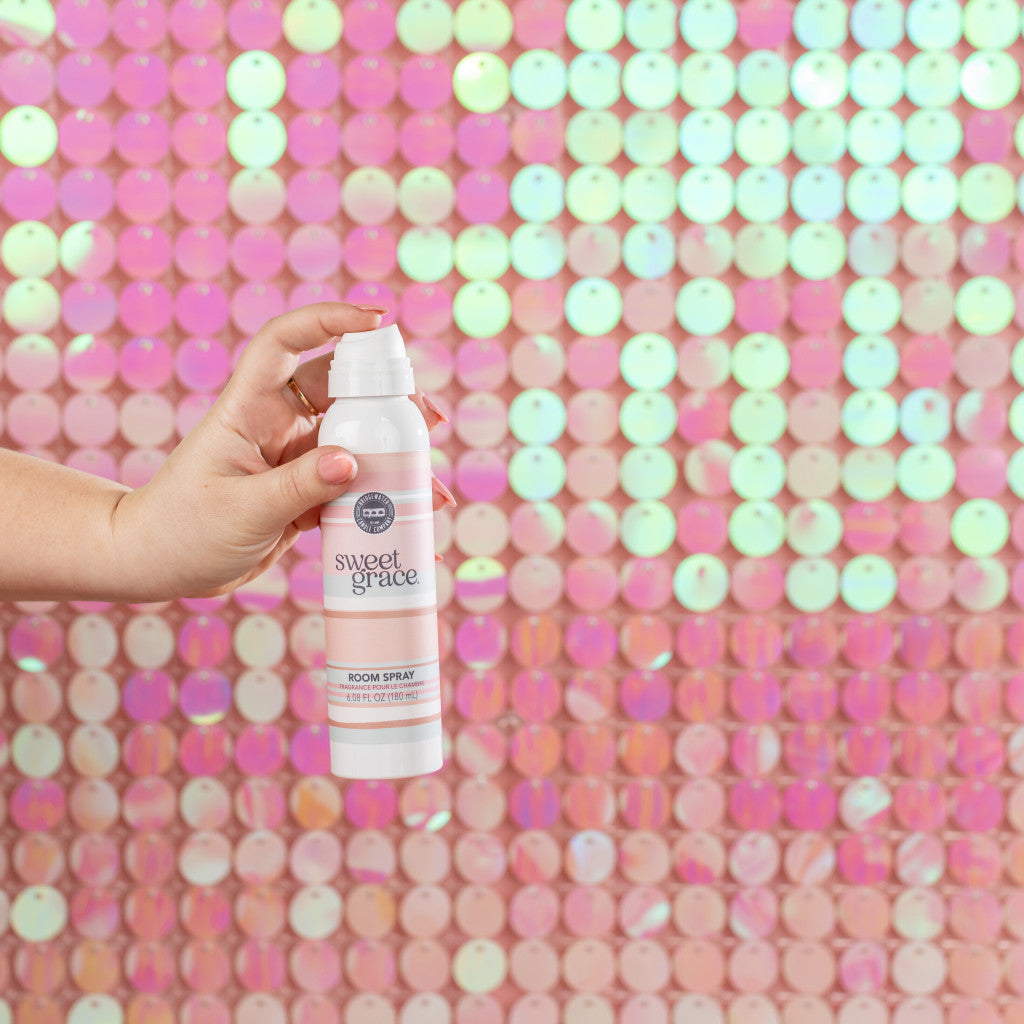 A hand holding a bottle of Sweet Grace room spray in front of a shimmering pink and white sequin backdrop.