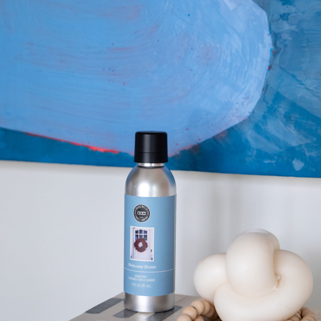 The Bridgewater Candle Company’s Welcome Home Non-Aerosol Room Spray, in a silver bottle with a blue label, sits beside a cream knotted candle and wooden beads beneath an abstract blue painting, creating a serene, eco-friendly vibe.