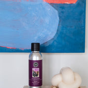 A purple bottle of Bridgewater Candle Companys Kiss in the Rain Non-Aerosol Room Spray rests on a wooden surface next to a cream knotted sculpture, with an abstract blue and red painting behind it.