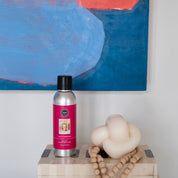 A silver bottle of Bridgewater Candle Companys Lets Celebrate Non-Aerosol Room Spray with a pink label sits on a patterned box beside a white knotted candle and wooden bead garland, with an abstract blue and red painting in the background.