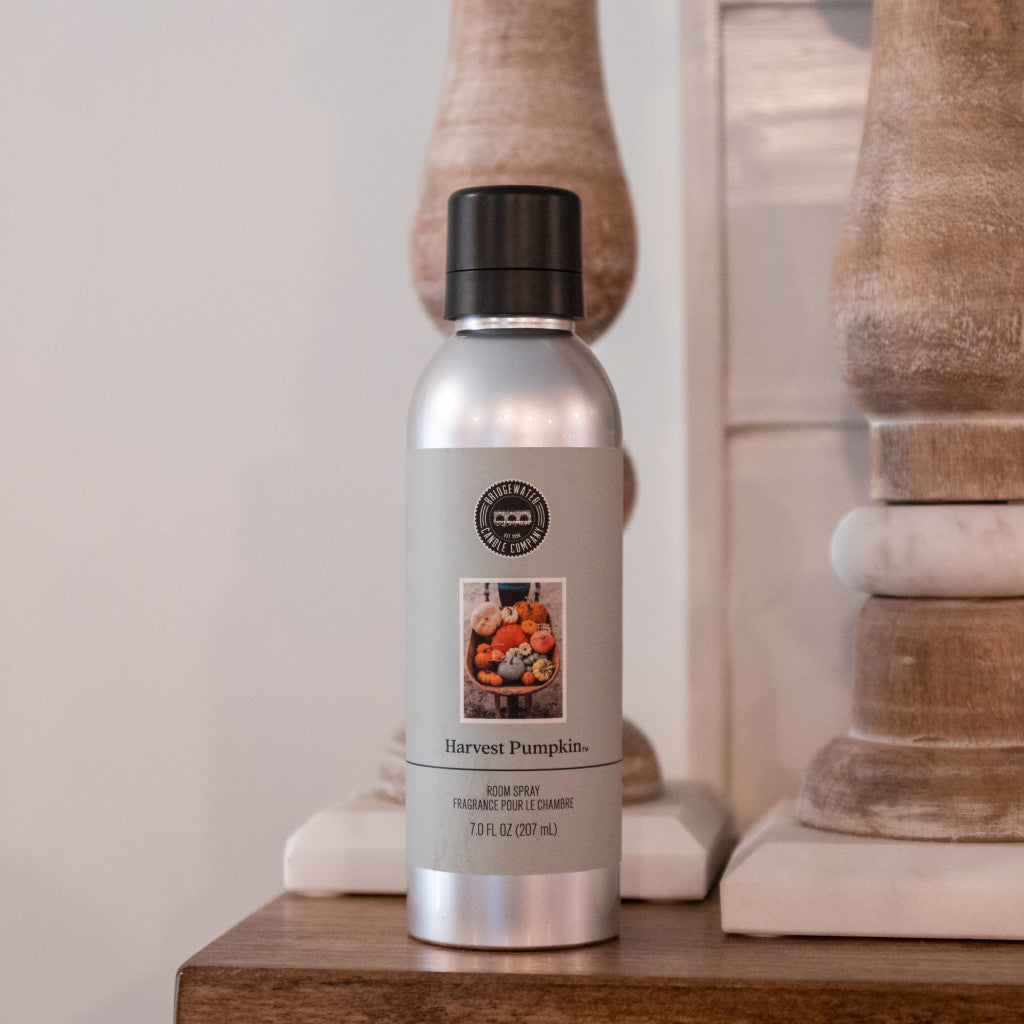 A silver canister of Bridgewater Candle Companys Harvest Pumpkin Non-Aerosol Room Spray with a black cap sits on wood, flanked by two decorative wooden objects in the background.
