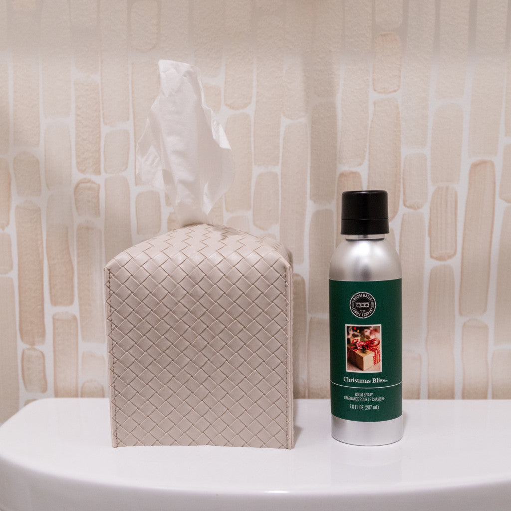 A textured tissue box and Bridgewater Candle Companys Christmas Bliss Non-Aerosol Room Spray rest side by side on a white toilet lid, with a beige patterned tiled wall in the background.