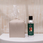 A textured tissue box and Bridgewater Candle Companys Christmas Bliss Non-Aerosol Room Spray rest side by side on a white toilet lid, with a beige patterned tiled wall in the background.