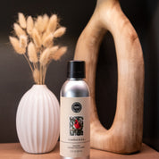 A silver Comfort & Joy Non-Aerosol Room Spray by Bridgewater Candle Company sits between a white ribbed vase with dried grass and a large wooden decorative piece on a wooden surface.