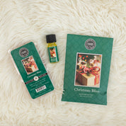 The Bridgewater Candle Company Christmas Bliss Flameless Bundle rests on a soft white surface, displaying a scented sachet, wax melt pack, and fragrance oil—all in festive holiday packaging for an inviting home fragrance.