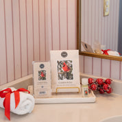 A bathroom counter displays the Bridgewater Candle Company Comfort & Joy Flameless Bundle on a white tray, alongside a small bottle of fragrance oil, a rolled white towel tied with red ribbon, and red berries by a gold-framed mirror.