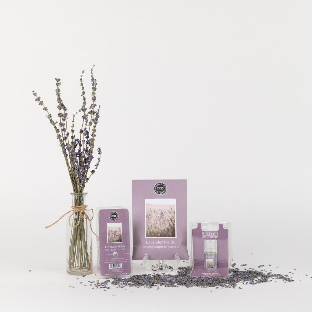 A glass jar with dried lavender sits beside the Bridgewater Candle Company Lavender Fields Flameless Bundle—featuring a card, sachet, and spray—displayed on a white surface with scattered lavender buds.