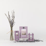 A glass jar with dried lavender sits beside the Bridgewater Candle Company Lavender Fields Flameless Bundle—featuring a card, sachet, and spray—displayed on a white surface with scattered lavender buds.