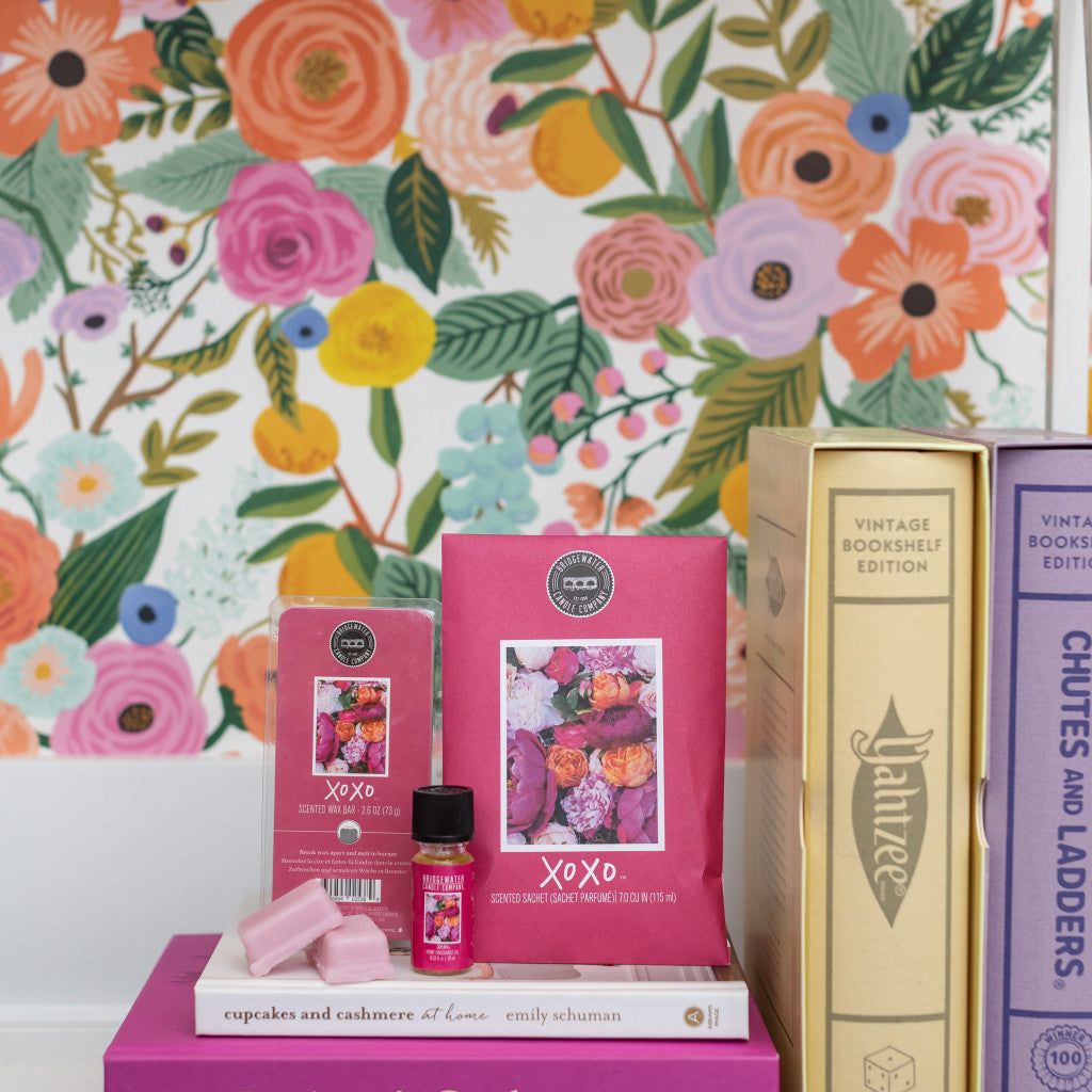 A colorful floral wallpaper frames books, a purple box, and Bridgewater Candle Companys XOXO Flameless Bundle products. A candle, soy-blend wax bar, fragrance oil, soap, and decorative books create a bright, cozy scene perfect for any room.