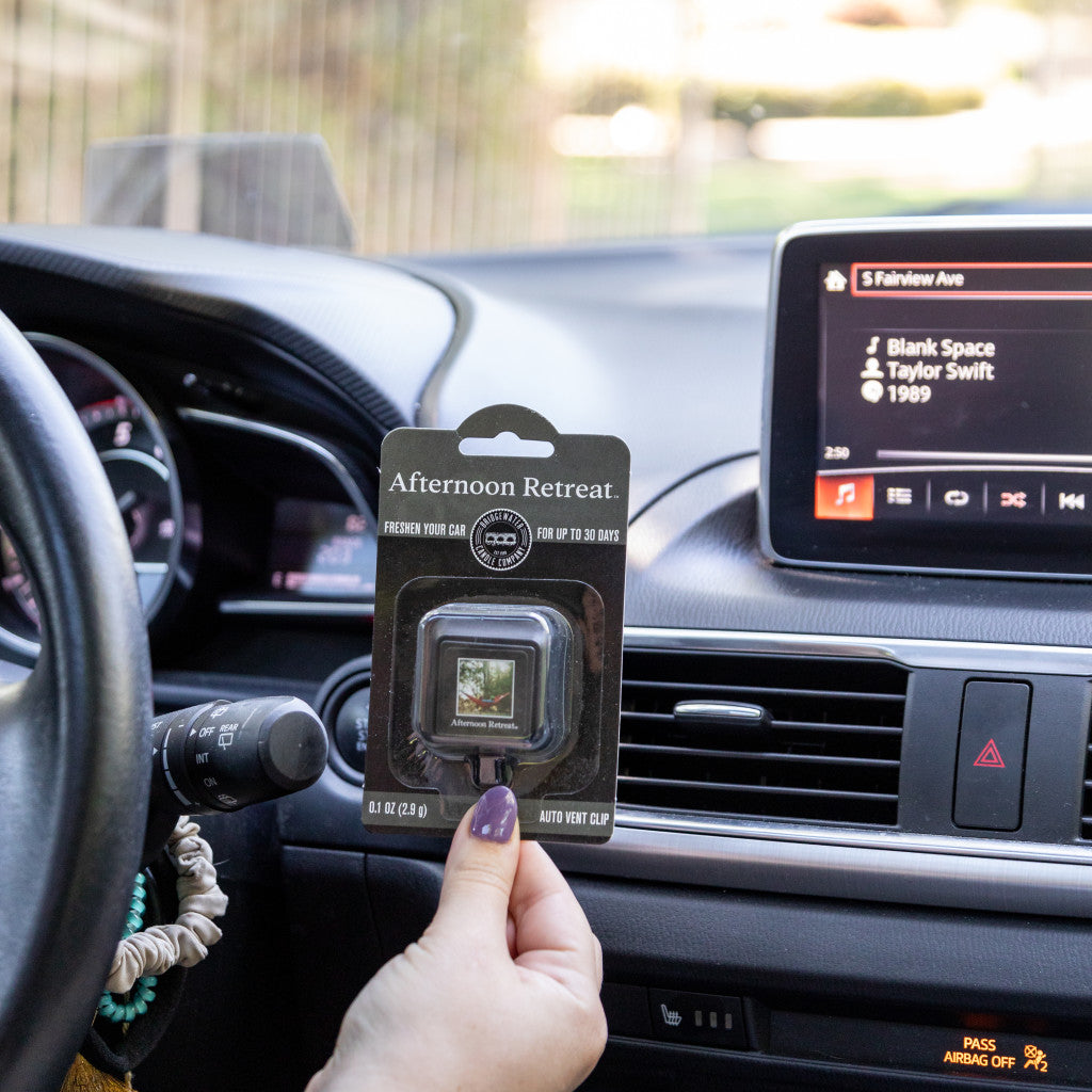 A hand holds a Bridgewater Candle Company Afternoon Retreat Auto Vent Clip in a car, with the dashboard showing Taylor Swifts Blank Space on the music screen, plus the steering wheel and air vent in view.