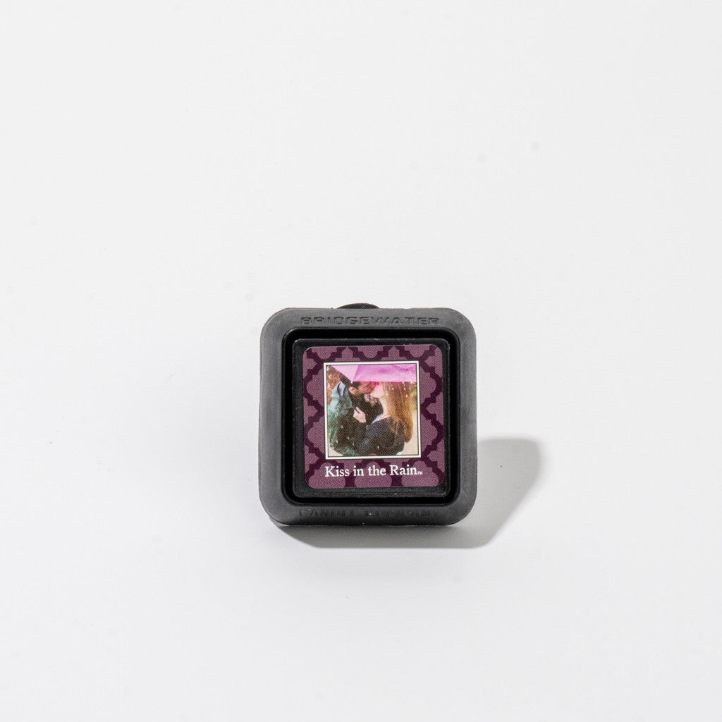 A small, square Kiss in the Rain Auto Vent Clip by Bridgewater Candle Company features two people kissing under an umbrella with the text Kiss in the Rain on a purple patterned background, centered on a plain white surface.