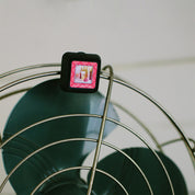 A small black Lets Celebrate Auto Vent Clip by Bridgewater Candle Company sits atop a metal wire fan guard, with large green blades in the background.