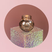 A small, round glass holder with a lit Bridgewater Candle Company Sweet Grace Collection Candle #008 sits on an iridescent pedestal against a pink background, filling the air with our best-selling fragrance.