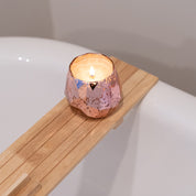 A lit Sweet Grace Collection Candle #011 by Bridgewater Candle Company in a faceted rose gold holder sits on a wooden bath tray above a white tub, filling the air with its best-selling fragrance.