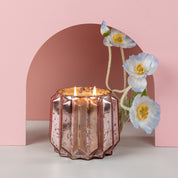 A Bridgewater Candle Company Sweet Grace Collection Candle #021 in a faceted rose gold mercury holder sits beneath a pink arch, beside delicate blue and white artificial flowers with yellow centers.