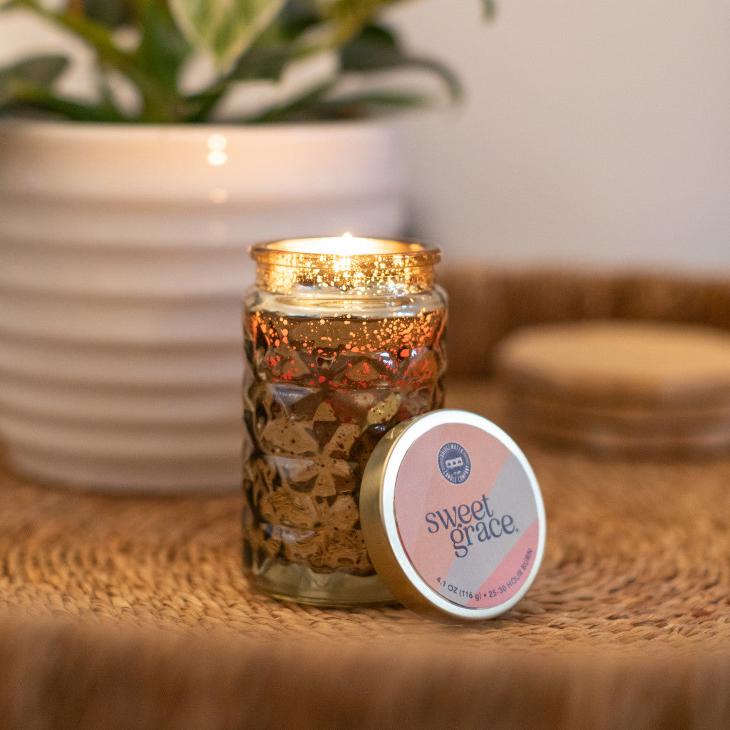 A lit Bridgewater Candle Company Sweet Grace Collection Candle #022 in a textured glass jar rests on a woven surface next to its matching tin, with a potted plant blurred in the background.