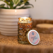 A lit Bridgewater Candle Company Sweet Grace Collection Candle #022 in a textured glass jar rests on a woven surface next to its matching tin, with a potted plant blurred in the background.