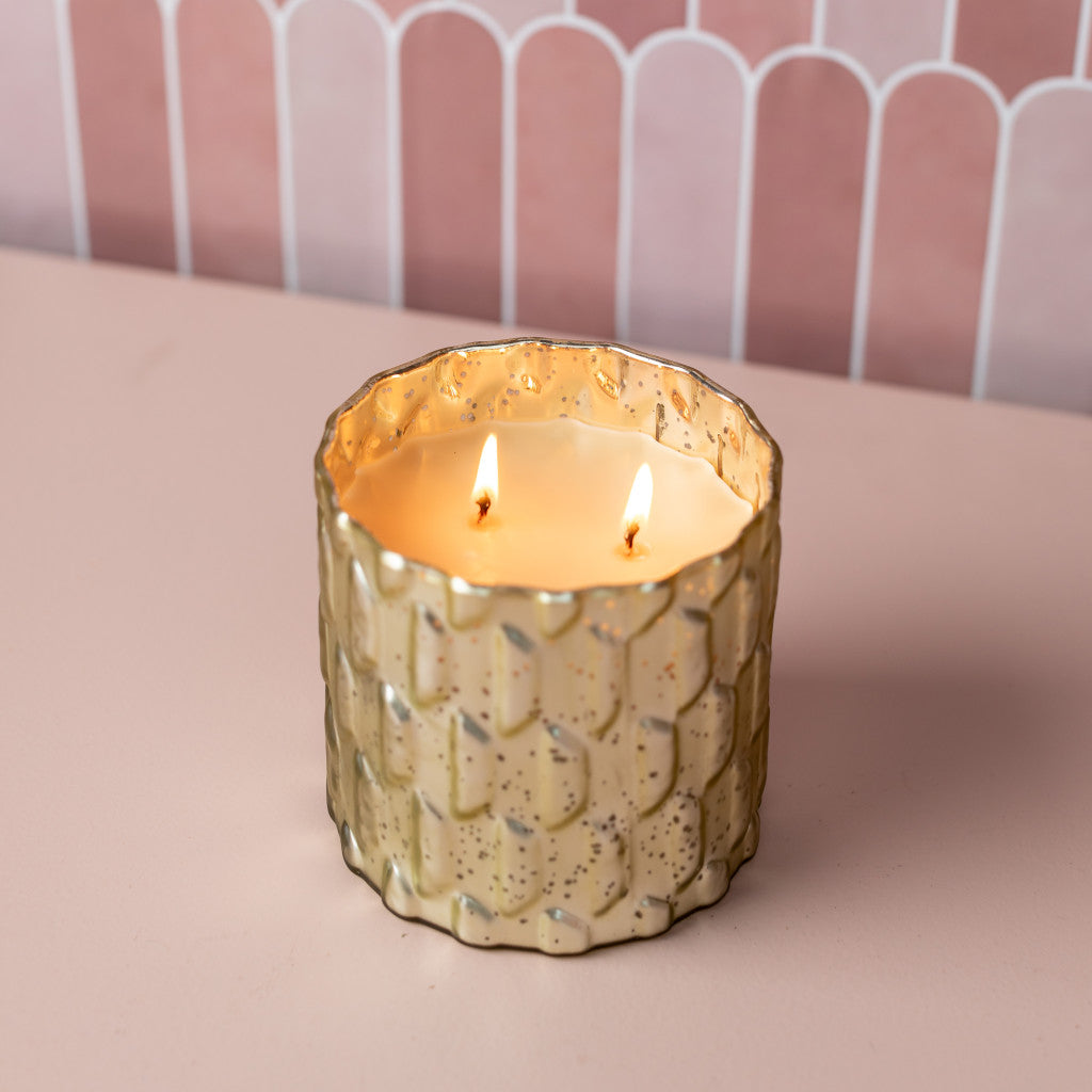 The Sweet Grace Collection Candle #035 by Bridgewater Candle Company glows with two flames in a textured gold glass holder atop a pale pink surface, set against a pink and white tiled wall, featuring its signature soy-blend wax.