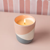A lit Bridgewater Candle Company Sweet Grace Collection Candle #041 with soy-blend wax sits in a ceramic holder featuring pink, white, and gray diagonal stripes on a light pink surface against a pink and white patterned background.