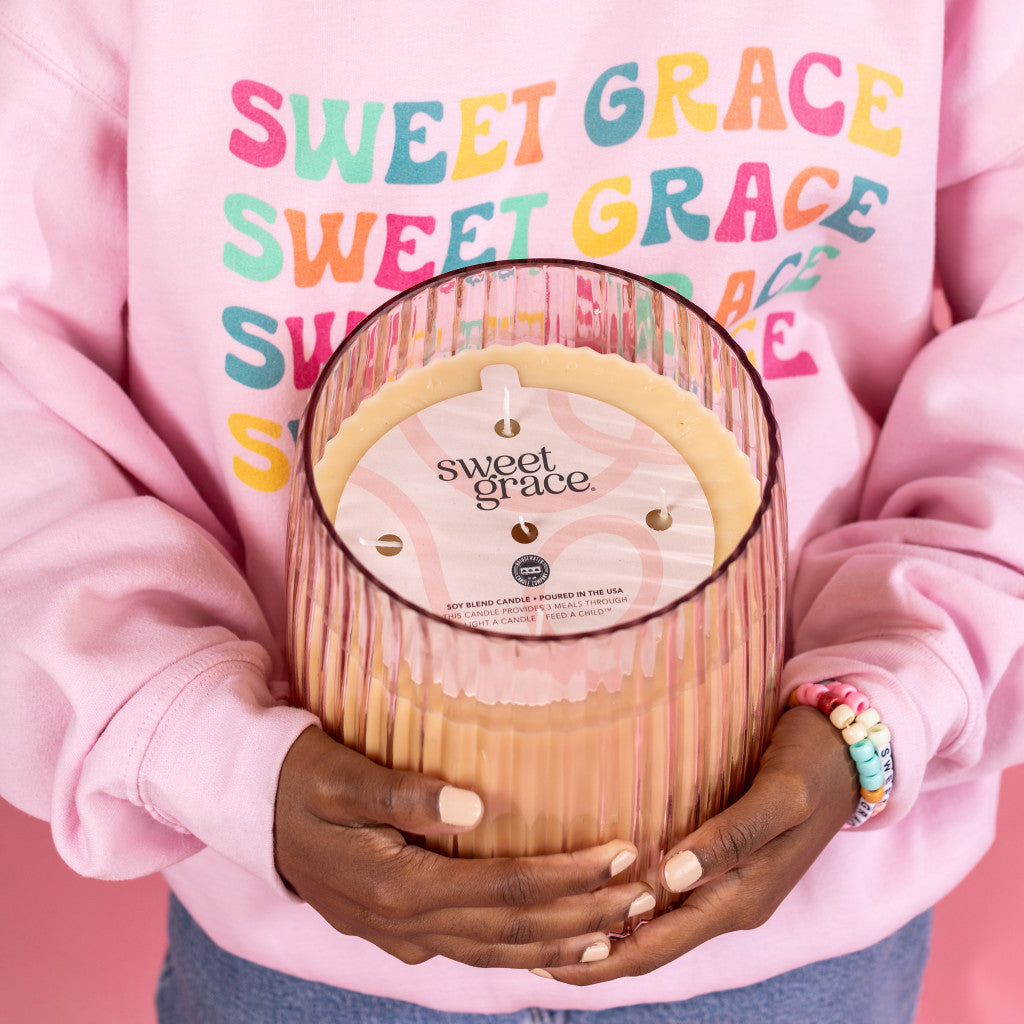 Wearing a pink sweatshirt with colorful SWEET GRACE text, a person holds the Bridgewater Candle Company Sweet Grace Collection Candle #043, a large soy-blend wax candle with five cotton wicks, standing against a pink background and wearing a beaded bracelet.