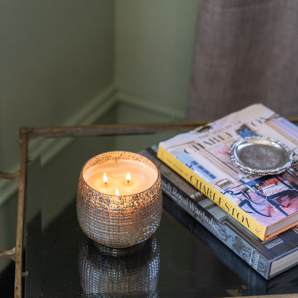 A lit Sweet Grace Collection Candle #051 by Bridgewater Candle Company, in a textured glass holder, glows on a glass table beside books and a metal dish, near a gray curtain and pale green wall.
