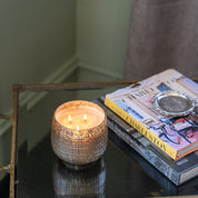 A lit Sweet Grace Collection Candle #051 by Bridgewater Candle Company, in a textured glass holder, glows on a glass table beside books and a metal dish, near a gray curtain and pale green wall.
