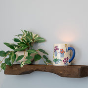 A small potted plant and a Bridgewater Candle Company Sweet Grace Collection Candle #054 in a white mug with colorful plant illustrations sit on a rustic wooden wall shelf against a light gray wall—perfect for any plant mom.