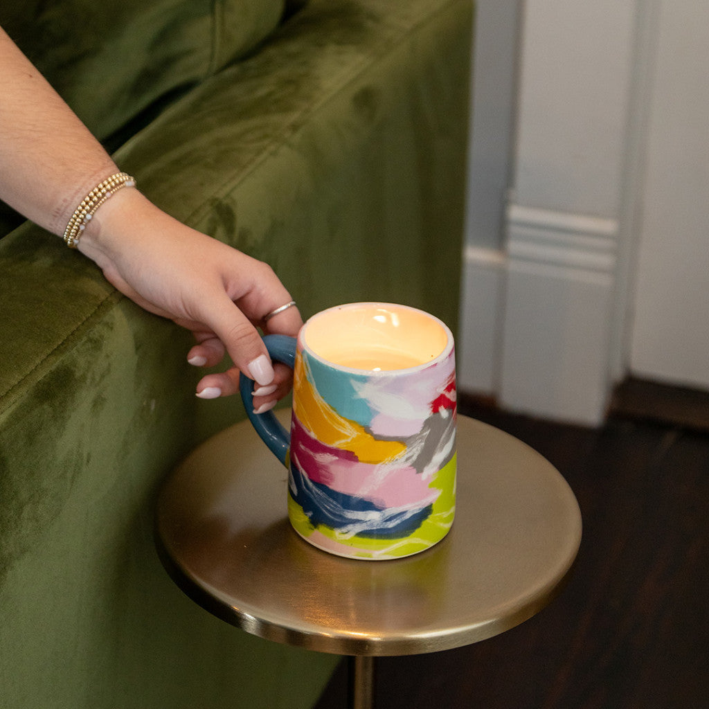 A hand with a beaded bracelet holds a colorful ceramic mug on a small gold table beside the Bridgewater Candle Company Sweet Grace Collection Candle #055, made from soy-blend wax, next to a green velvet couch.