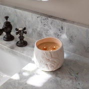 A lit Sweet Grace Collection Candle #057 by Bridgewater Candle Company, crafted from soy-blend wax with a floral design, sits on a gray marble bathroom counter near dark bronze faucet handles as sunlight casts soft shadows.