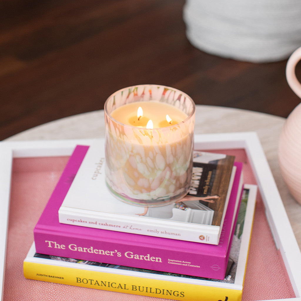 A lit Bridgewater Candle Company Sweet Grace Collection Candle #058, made with soy-blend wax and multiple wicks, sits atop colorful books—The Gardener’s Garden and Botanical Buildings—on a white tray on the coffee table.