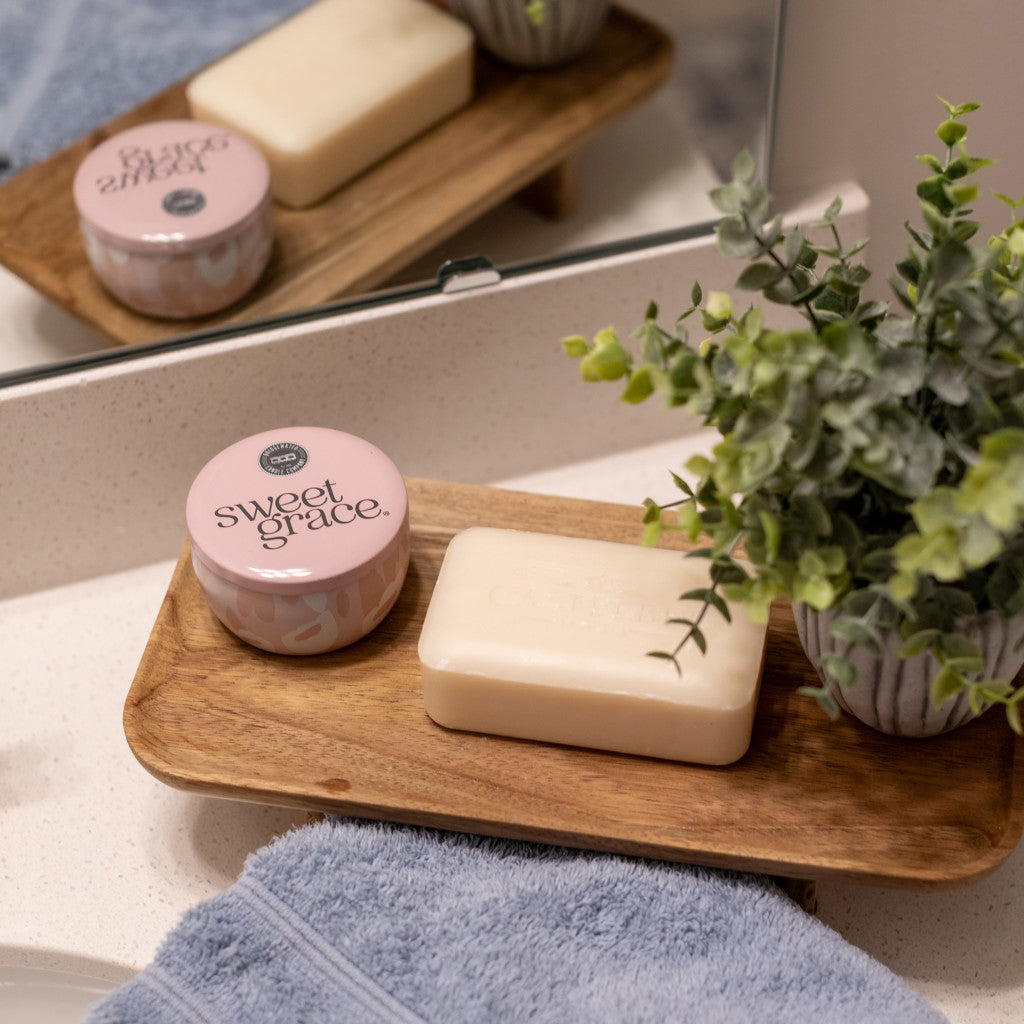 A wooden tray on the bathroom counter holds a bar of soap, a small potted plant, and a Bridgewater Candle Company Sweet Grace Collection Candle #059, with a blue towel nearby and the mirror reflecting the serene scene.