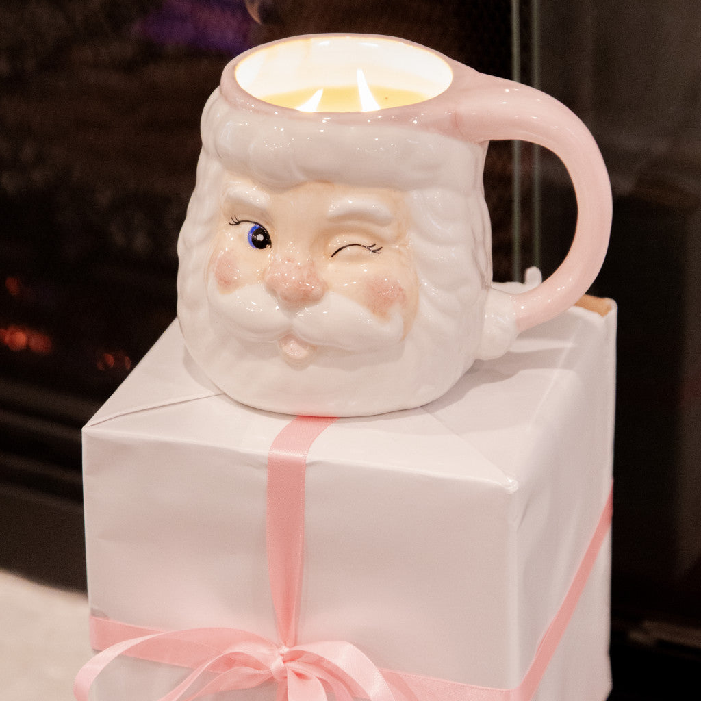 The Bridgewater Candle Company Sweet Grace Santa Mug, a white mug shaped like a winking Santa with a pink handle, rests on a white gift box tied with a pink ribbon.