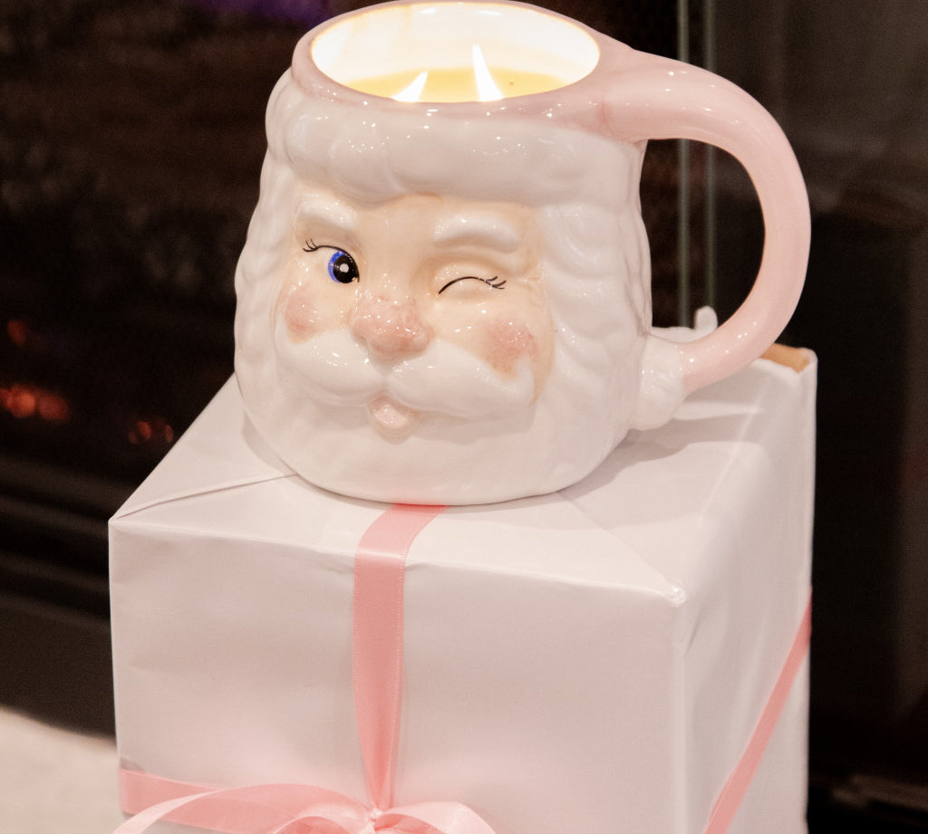 The Bridgewater Candle Company Sweet Grace Santa Mug, a white mug shaped like a winking Santa with a pink handle, rests on a white gift box tied with a pink ribbon.