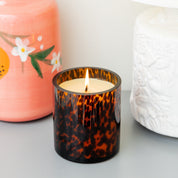A lit Bridgewater Candle Company Sweet Grace Collection Candle #064 in a brown tortoise glass holder sits on a gray surface, with a pink floral vase and a white textured vase in the background.
