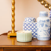 A Sweet Grace Collection Candle #065 by Bridgewater Candle Company, in a green ribbed vessel, sits on a wooden table beside two blue and white ceramic jars and a gold twisted-base lamp.